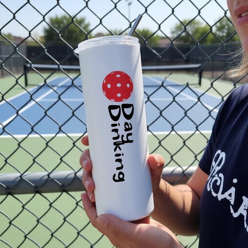 May include: A white cylindrical tumbler with a clear lid and a metal straw. The tumbler features a red pickleball graphic above the black text "Day Dinking" printed vertically. The design is centered on the smooth surface of the drinkware.