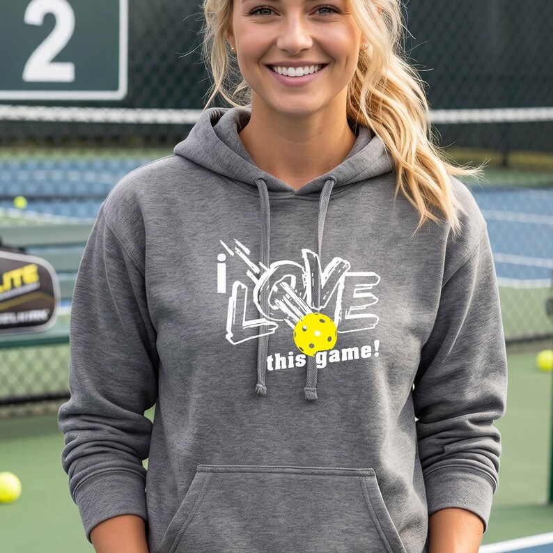 A grey hooded sweatshirt featuring a white graphic design that reads "I LOVE this game!" with a yellow pickleball replacing the letter "O". The hoodie includes a matching drawstring cord at the neckline and long sleeves.