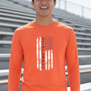 A bright orange long-sleeve shirt featuring a vertical graphic design of a distressed American flag with the word PICKLEBALL printed in white capital letters running vertically through the center of the flag stripes.