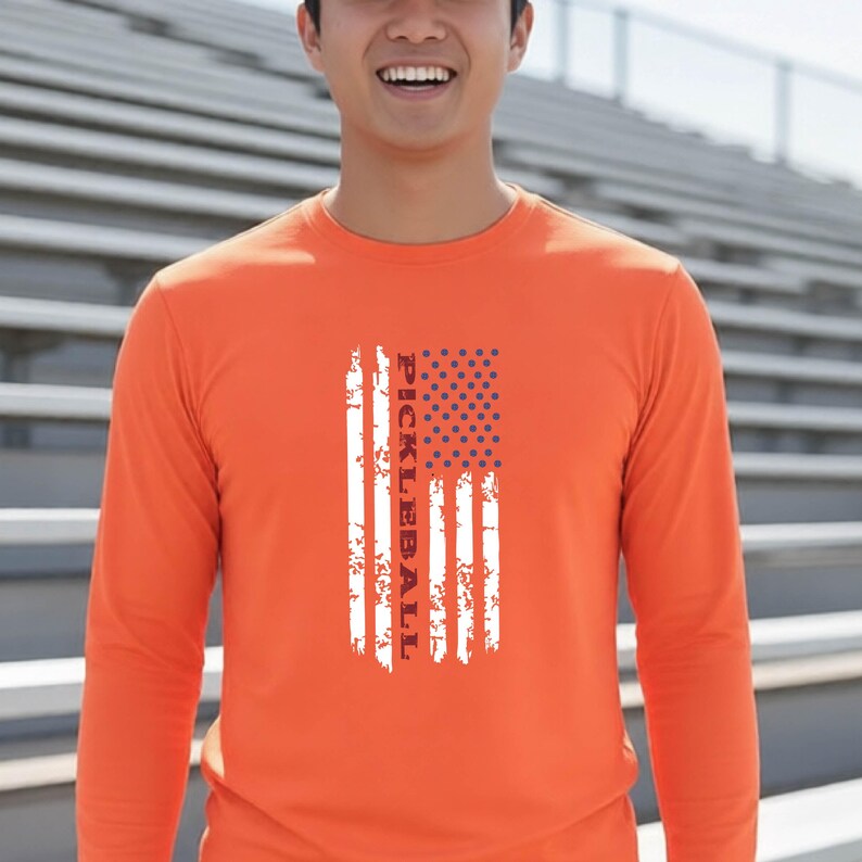 A bright orange long-sleeve shirt featuring a vertical graphic design of a distressed American flag with the word PICKLEBALL printed in white capital letters running vertically through the center of the flag stripes.