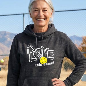 A charcoal gray long-sleeve hooded sweatshirt featuring a white graphic print that reads I LOVE this game! with a yellow pickleball replacing the letter O. The hoodie includes a front kangaroo pocket and matching drawstring cords.