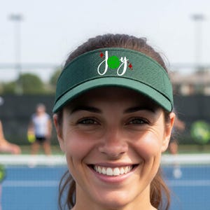 Könnte beinhalten: Ein dunkelgrünes Visier mit dem Wort Joy in weißer Schreibschrift. Der Buchstabe O ist durch eine grüne Pickleball-Grafik ersetzt. Kleine rote Rautenformen befinden sich oberhalb und rechts neben dem Text auf der Vorderseite des Visiers.