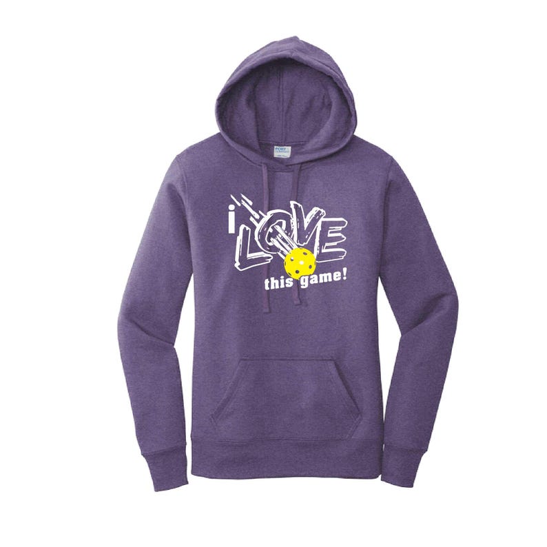 K&ouml;nnte beinhalten: Lila Kapuzenpullover mit Fronttasche und Kordelzugkapuze. Auf der Vorderseite steht in Wei&szlig; der Text "i LOVE this game!" mit einem gelben Pickleball. Ein l&auml;ssiges, bequemes Kleidungsst&uuml;ck.