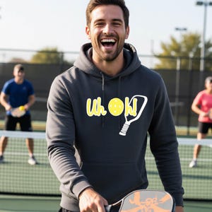 A dark gray hooded sweatshirt featuring a white graphic print on the chest. The design includes the text "Uh oh!" in a playful font, a yellow pickleball with small dots, and a white outline of a pickleball paddle with a grip.
