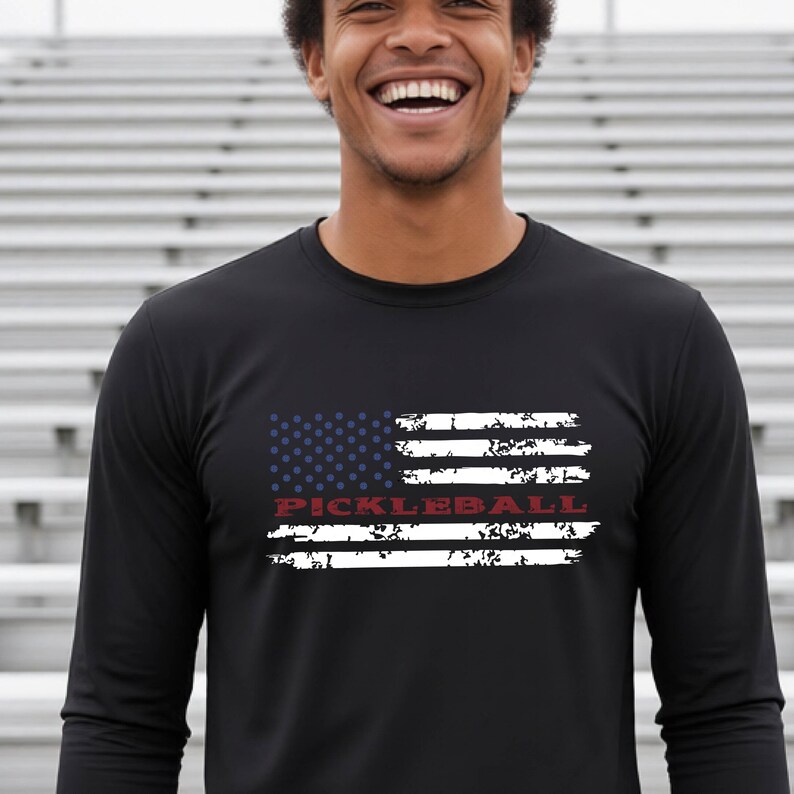 A black long-sleeve shirt featuring a distressed graphic of the American flag in white and blue, with the word PICKLEBALL printed in red across the center of the flag design. The shirt has a crew neckline and long sleeves.