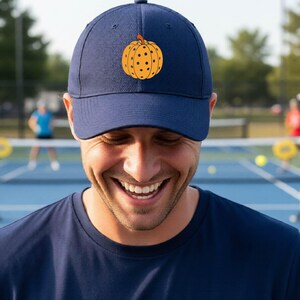 Könnte beinhalten: Eine marineblaue Baseballkappe mit einer mittigen Grafik eines orangefarbenen Kürbisses mit schwarzen Punkten. Die Kappe verfügt über eine strukturierte Krone, einen gebogenen Schirm und genähte Ösen an den oberen Paneelen.