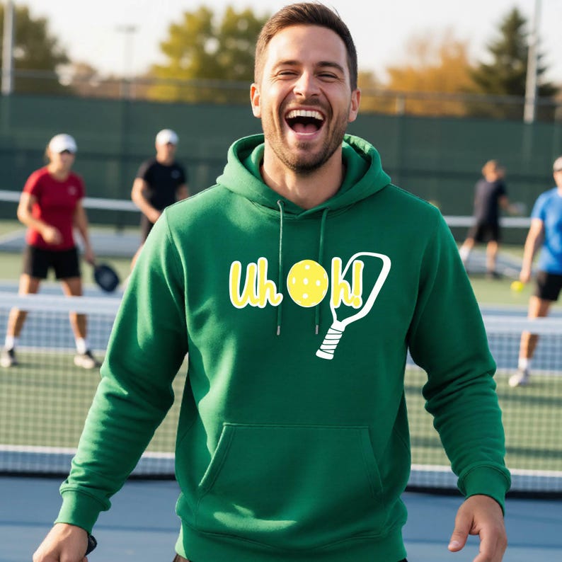 A forest green hooded sweatshirt featuring a front kangaroo pocket and a graphic print on the chest. The design includes the yellow text "Uh oh!" alongside a yellow pickleball and a white outline of a pickleball paddle.