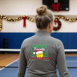 May include: A grey long-sleeved zip-up jacket featuring a graphic on the back. The design includes the text "SANTA'S FAVORITE PICKLEBALLER" in green and red, with a yellow pickleball wearing a red and white Santa hat between two green trees.