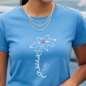 Light blue t-shirt featuring a white floral design with a pink center and the word "Pickleball" in a script font. The shirt is made of a soft material and is suitable for casual wear.