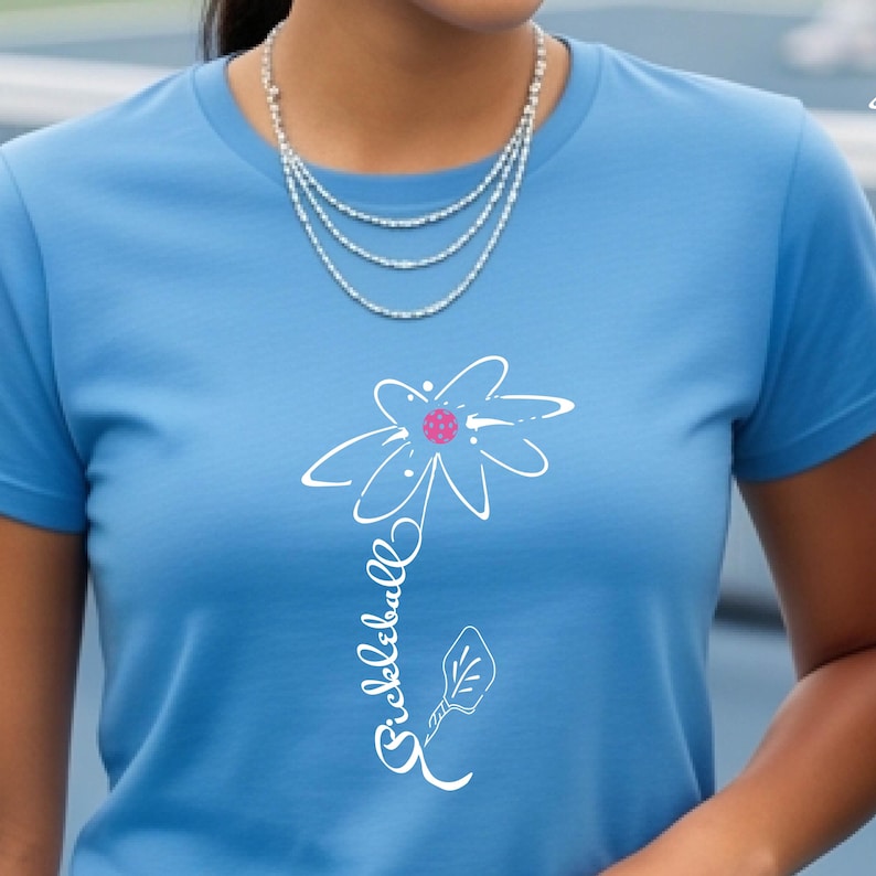 Light blue t-shirt featuring a white floral design with a pink center and the word "Pickleball" in a script font. The shirt is made of a soft material and is suitable for casual wear.