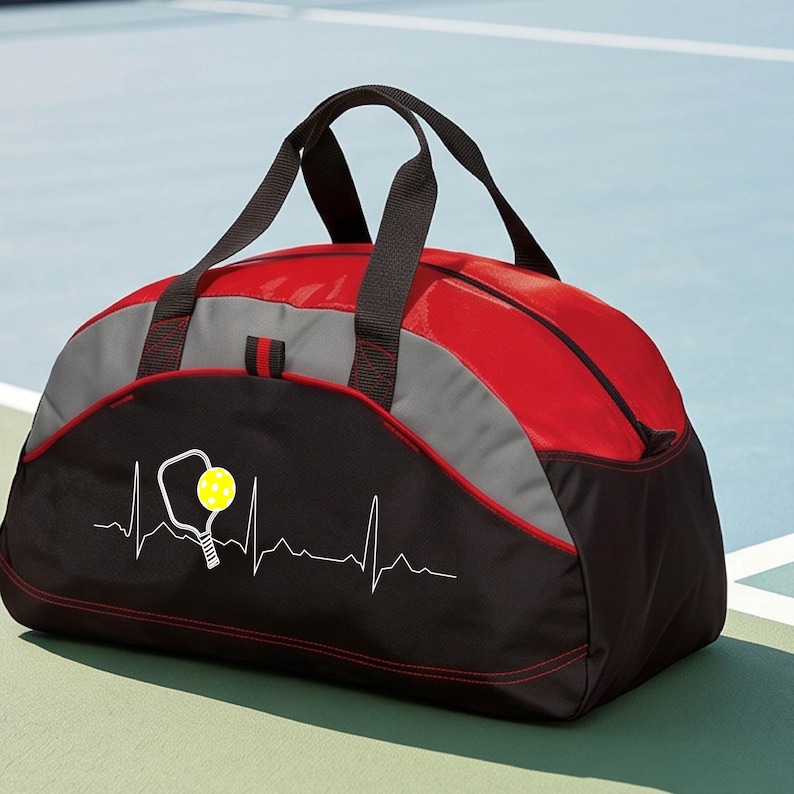 May include: A red, grey and black duffel bag featuring a white heartbeat EKG line graphic with a white pickleball paddle and a yellow pickleball. The bag has two black top handles, a zippered main compartment, and red stitching accents.