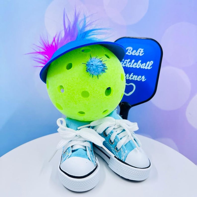 May include: A green pickleball with a blue hat and a blue and white feather on top. The pickleball is wearing blue sneakers and is holding a blue paddle that says "Best Pickleball Partner".