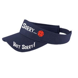 May include: A navy blue visor with white text that reads "SORRY... NOT SORRY!" and a red and white pickleball graphic.