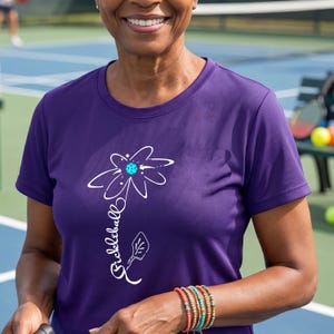 A purple t-shirt with a white floral design and the word "Pickleball" in script. The flower has a blue center. The shirt is short-sleeved and made of a textured fabric. The person is wearing a bracelet with colorful beads.