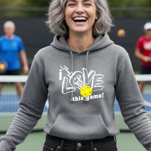 A heather gray long-sleeve hooded sweatshirt featuring a white graphic print that reads I LOVE this game! with a yellow pickleball replacing the letter O. The hoodie includes a front kangaroo pocket and matching gray drawstrings.