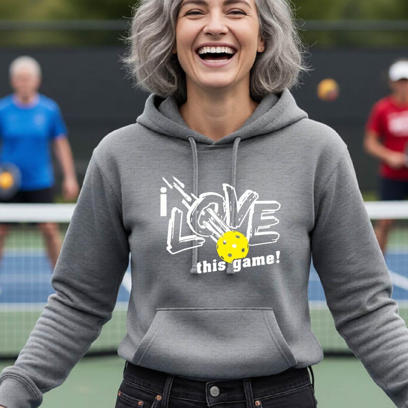A heather gray long-sleeve hooded sweatshirt featuring a white graphic print that reads I LOVE this game! with a yellow pickleball replacing the letter O. The hoodie includes a front kangaroo pocket and matching gray drawstrings.
