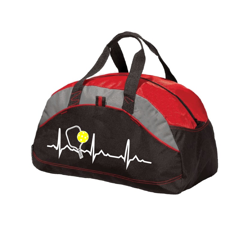May include: A black, red, and grey duffel bag featuring a white heartbeat line graphic that incorporates a pickleball paddle and a yellow pickleball. The bag has two black fabric handles and a zippered main compartment for storage.
