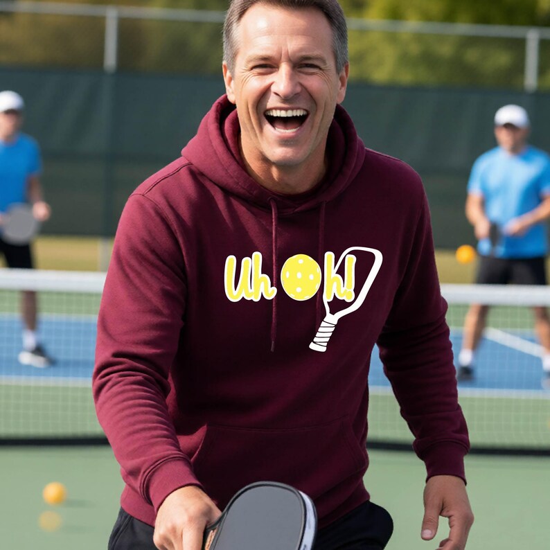 A maroon long-sleeve hooded sweatshirt featuring a graphic print on the chest with the yellow text "Uh oh!", a yellow pickleball, and a white pickleball paddle outline. The hoodie includes a front kangaroo pocket and matching drawstrings.