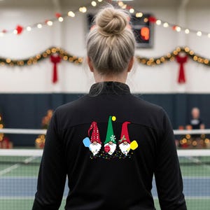 Puede incluir: Una chaqueta deportiva negra de manga larga con un gráfico en la espalda de tres gnomos con gorros festivos. Los gnomos sostienen paletas de pickleball y están rodeados de luces de colores. La chaqueta tiene un cuello alto y mangas largas.