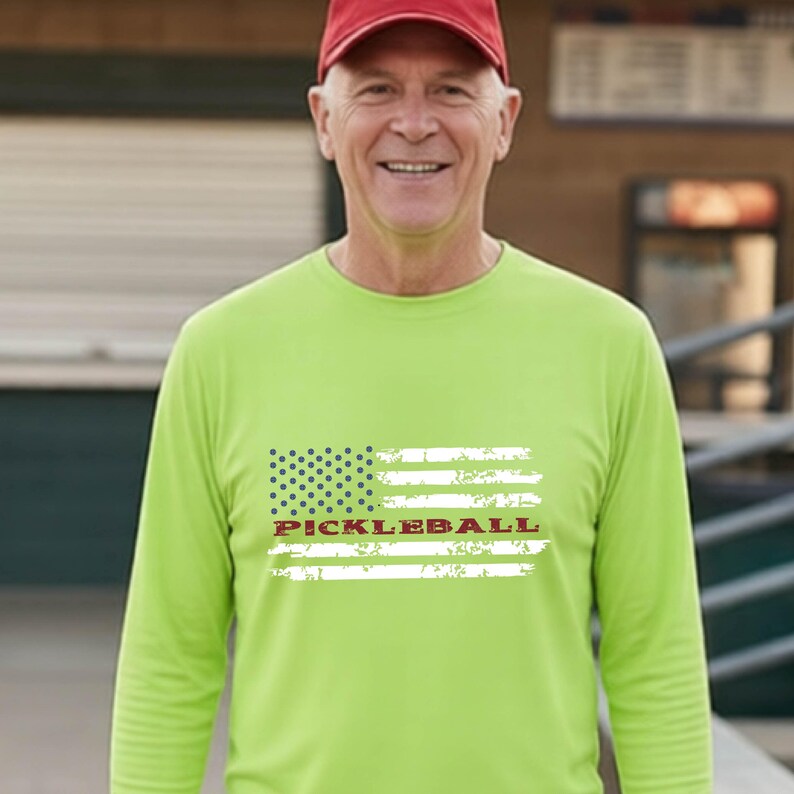 A long-sleeve lime green shirt featuring a white distressed American flag graphic with the word PICKLEBALL printed in red across the center. The shirt has a crew neckline and is worn with a solid red baseball cap.