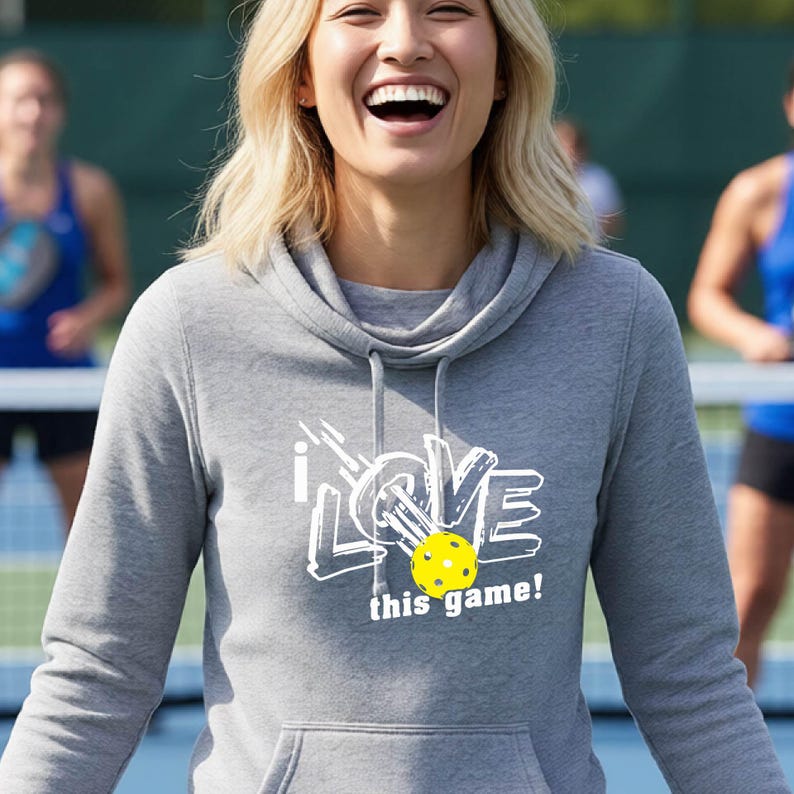 A heather gray long-sleeve hoodie with a cowl neckline and a front kangaroo pocket. The chest features a white graphic design with the text "i LOVE this game!" and a bright yellow pickleball with small circular holes.