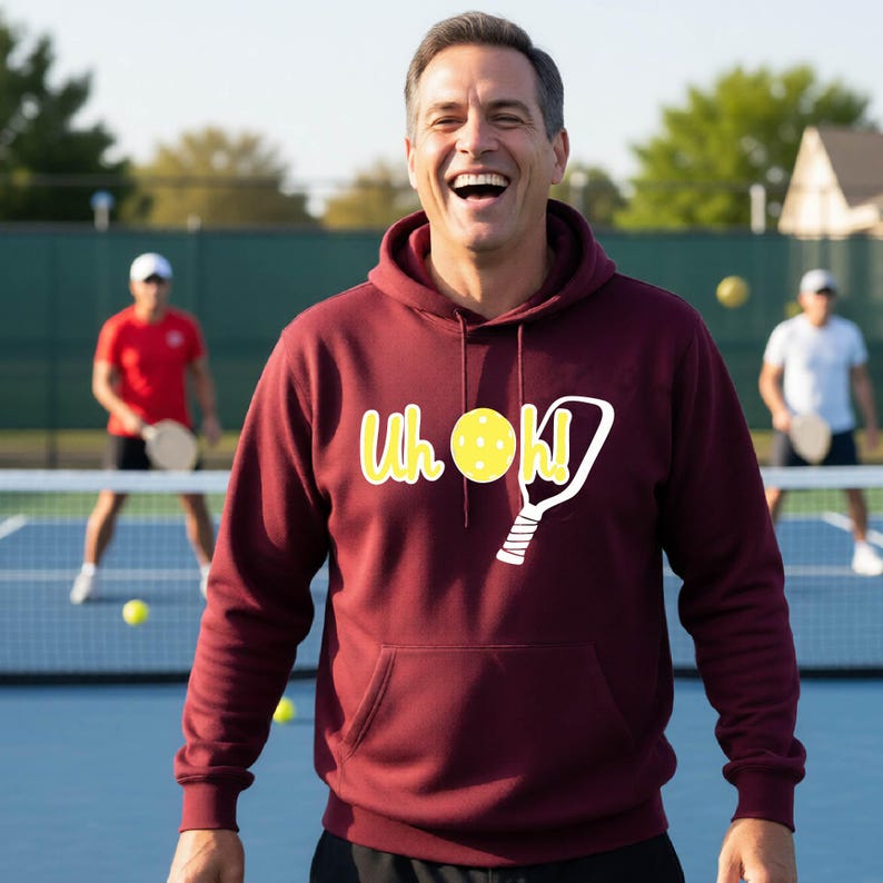 A maroon hooded sweatshirt featuring a graphic print on the chest. The design includes the yellow text "Uh oh!" with a yellow pickleball replacing the letter "o" and a white outline of a pickleball paddle next to the text.