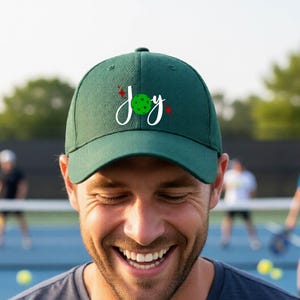 Könnte beinhalten: Eine dunkelgrüne Baseballkappe mit dem weißen Schriftzug "Joy" auf der Vorderseite. Der Buchstabe "o" ist durch einen grünen Pickleball mit kleinen schwarzen Punkten ersetzt. Zwei kleine rote vierzackige Sterne befinden sich neben dem Text.