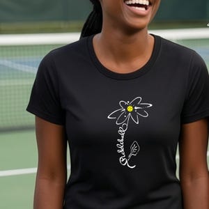 Black t-shirt with a white and yellow daisy design. The daisy has a yellow center and the word "pickleball" is written in a cursive font along the stem. The shirt has a crew neck.