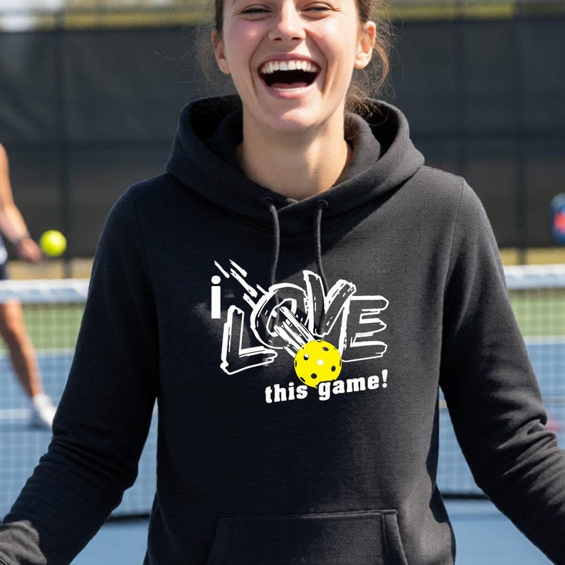 A black hooded sweatshirt featuring a white graphic design with the text "i LOVE this game!" and a yellow pickleball with black holes integrated into the lettering. The hoodie includes a front kangaroo pocket and black drawstrings.