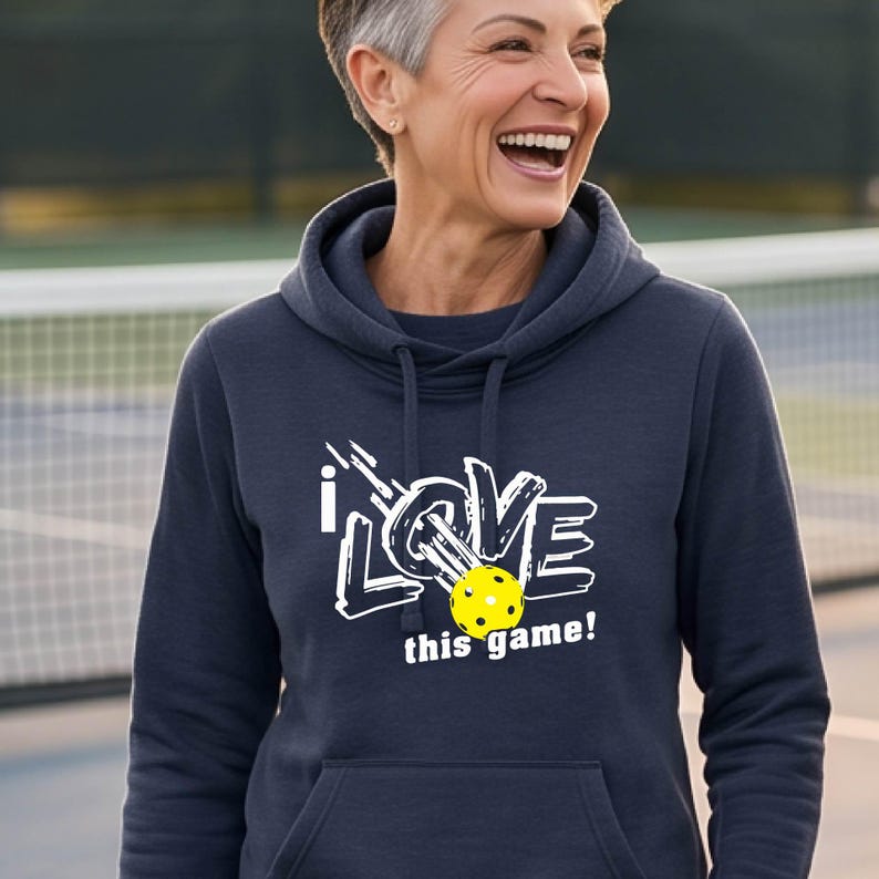 A navy blue hooded sweatshirt featuring a white graphic design that reads "I LOVE this game!" with a yellow pickleball replacing the letter "O". The hoodie includes a matching drawstring cord at the neckline and long sleeves.