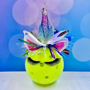 May include: A yellow pickleball ornament decorated with a unicorn theme. It has a sparkly purple and blue crown, white and pink bow, and black eyelashes.