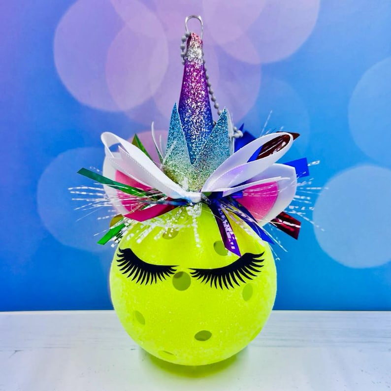 May include: A yellow pickleball ornament decorated with a unicorn theme. It has a sparkly purple and blue crown, white and pink bow, and black eyelashes.