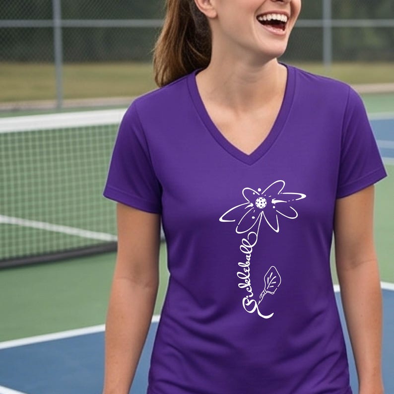 A purple V-neck t-shirt featuring a white floral design with a pickleball in the center. The word "pickleball" is written in cursive along the stem of the flower. The shirt is made of a lightweight material.