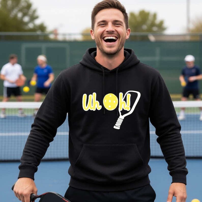 A black hooded sweatshirt featuring a front kangaroo pocket and a graphic print on the chest. The design includes the yellow text "Uh oh!" alongside a yellow pickleball and a white outline of a pickleball paddle.