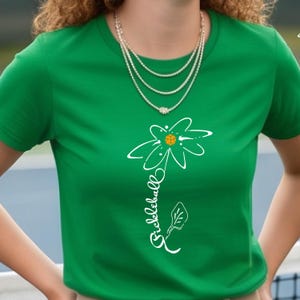 A green t-shirt featuring a white floral design with a pickleball in the center. The word "Pickleball" is written vertically in white script. A multi-strand necklace is worn around the neck.