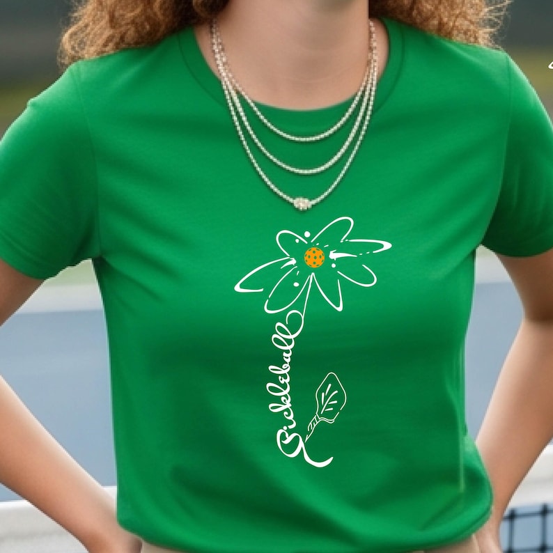 A green t-shirt featuring a white floral design with a pickleball in the center. The word "Pickleball" is written vertically in white script. A multi-strand necklace is worn around the neck.