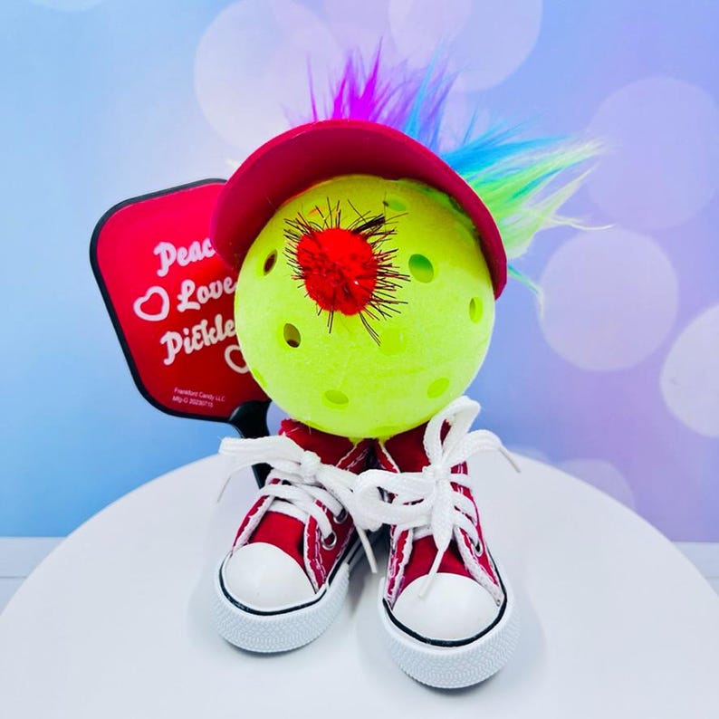 May include: A green pickleball with a red pom-pom on top, wearing a red hat with a rainbow feather mohawk and red and white sneakers. The pickleball is holding a red paddle with the words "Peace Love Pickleball" written on it.
