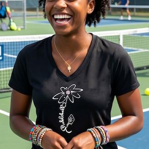 Black V-neck t-shirt with a white floral design and the word "Pickleball" in script. A gold necklace with a pickleball paddle pendant is worn. Colorful bracelets adorn the wrist. The background shows a pickleball court.