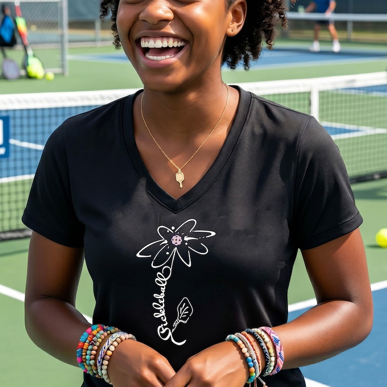 Black V-neck t-shirt with a white floral design and the word "Pickleball" in script. A gold necklace with a pickleball paddle pendant is worn. Colorful bracelets adorn the wrist. The background shows a pickleball court.