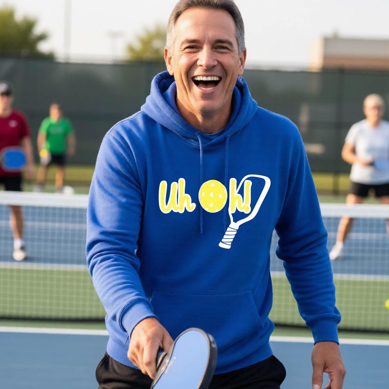 A royal blue hooded sweatshirt featuring a white graphic print that reads "Uh Oh!" with a yellow pickleball replacing the letter "O" and a white pickleball paddle illustration next to the text, worn on a pickleball court.