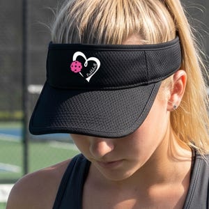May include: A black athletic visor made of a textured mesh fabric. The front of the visor features a white heart-shaped graphic outline with a pink pickleball positioned on the left side and the word Pickleball written in white script inside.