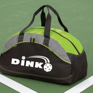 K&ouml;nnte beinhalten: Eine schwarze und lindgr&uuml;ne Sporttasche mit schwarzen Tragegriffen und einem grauen Akzentstreifen. Auf der Vorderseite befindet sich der wei&szlig;e Schriftzug DINK mit einer Grafik eines Pickleballs und Bewegungslinien sowie lindgr&uuml;nen N&auml;hten.
