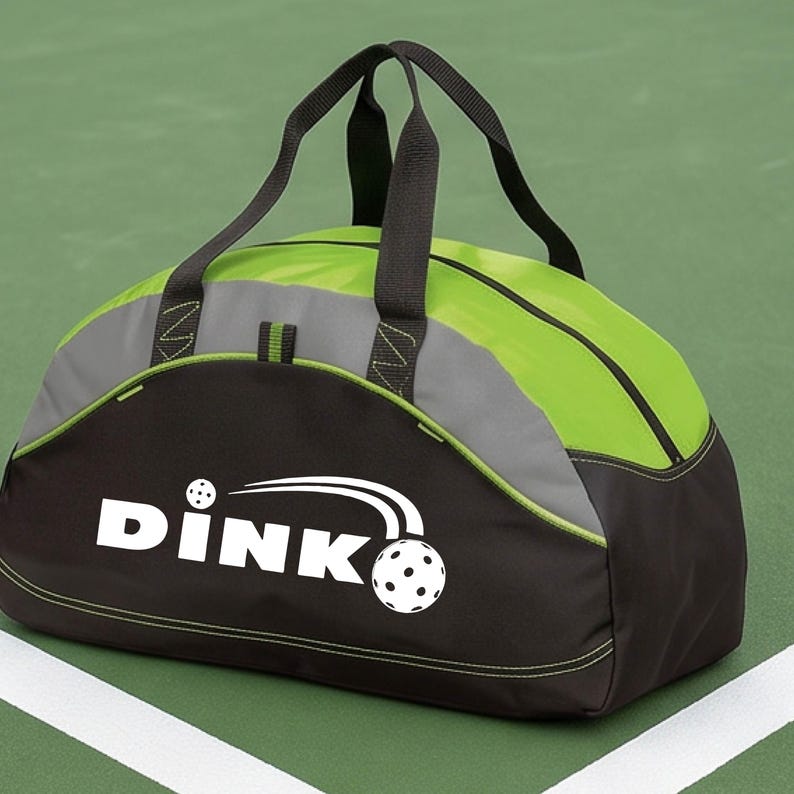 K&ouml;nnte beinhalten: Eine schwarze und lindgr&uuml;ne Sporttasche mit schwarzen Tragegriffen und einem grauen Akzentstreifen. Auf der Vorderseite befindet sich der wei&szlig;e Schriftzug DINK mit einer Grafik eines Pickleballs und Bewegungslinien sowie lindgr&uuml;nen N&auml;hten.