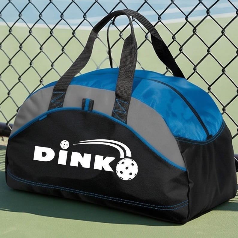 K&ouml;nnte beinhalten: Eine Sporttasche in Blau, Grau und Schwarz mit zwei schwarzen Tragegriffen. Auf der Vorderseite befindet sich der wei&szlig;e Schriftzug DINK sowie eine Grafik eines Pickleballs mit Bewegungslinien &uuml;ber dem Buchstaben K, aufgedruckt auf dem schwarzen Teil.