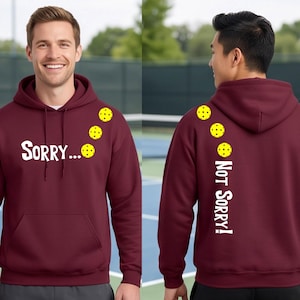 May include: Maroon hoodie with the text "Sorry..." on the front and "Not Sorry!" on the back. The hoodie features yellow pickleball graphics. The hoodie has a front pocket and a hood.