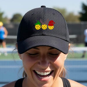 Könnte beinhalten: Eine schwarze Baseballkappe mit einer Grafik von drei gelben Pickleballs in einer Reihe. Der erste Ball trägt eine grüne Elfenmütze, der zweite hat ein braunes Rentiergeweih und der dritte trägt eine rote Weihnachtsmütze mit weißem Bommel.
