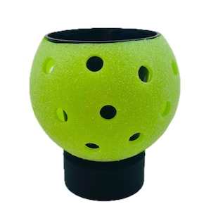 May include: A bright neon green pickleball with a textured surface and multiple circular holes, mounted on a solid black cylindrical base. The ball features a hollow interior and a smooth black rim at the top opening, creating a functional holder.