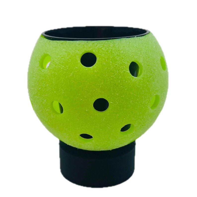 May include: A bright neon green pickleball with a textured surface and multiple circular holes, mounted on a solid black cylindrical base. The ball features a hollow interior and a smooth black rim at the top opening, creating a functional holder.