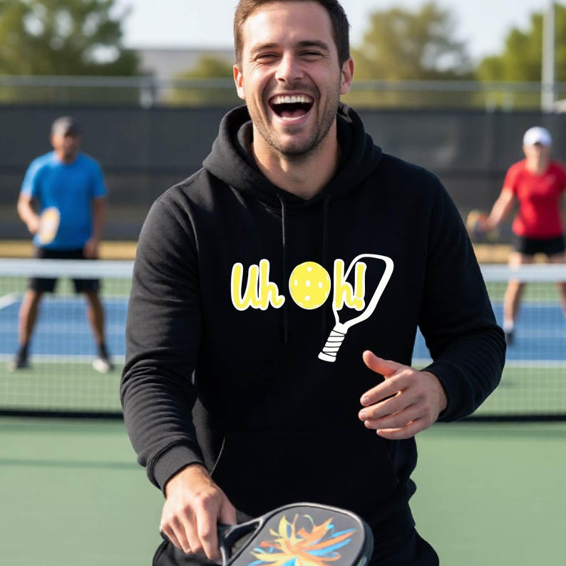 A black hooded sweatshirt featuring a graphic print on the chest with the yellow text Uh Oh! accompanied by a white pickleball paddle icon and a yellow pickleball. The hoodie has long sleeves, a drawstring hood, and a front kangaroo pocket.