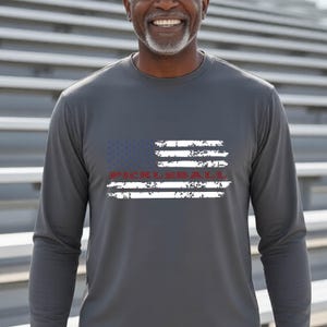 A charcoal gray long-sleeve shirt featuring a distressed American flag graphic in white and blue. The word PICKLEBALL is printed in red across the center of the flag design, which has a weathered, textured appearance on the fabric.